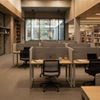 Architecturally stunning university library interior with exposed concrete beams and high wooden ceilings. The space features arranged acoustic modular study carrels with wood finishes and grey felt dividers, offering individual workstations with task lighting, laptops, and plants.