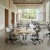 Bright modern university open study area featuring mobile desks and chairs arranged in flexible configurations with students working on laptops near large windows.