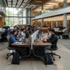 A modern university library study area with students working at back-to-back computer workstations, featuring a light gray privacy screen, integrated power outlets, and large windows overlooking a campus green.
