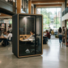 A student in a gray acoustic study pod with a wooden frame, studying with a laptop and books in a modern university library common area
